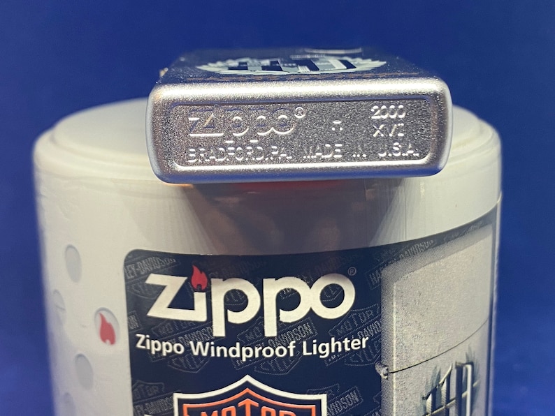 Puede incluir: Un encendedor Zippo Windproof plateado con el texto "Zippo" y "Bradford, PA. Made in U.S.A." impreso en la parte inferior. El encendedor est&aacute; sobre un recipiente blanco con un logotipo de Harley-Davidson.