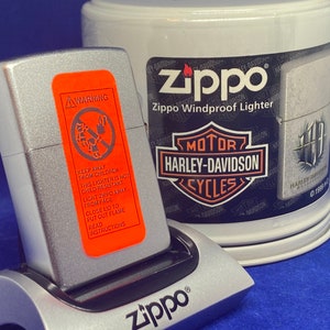 Puede incluir: Un encendedor Zippo plateado con una etiqueta de advertencia naranja y un soporte de carga plateado. La etiqueta dice "Mantener alejado de los ni&ntilde;os" y otras instrucciones de seguridad. Un recipiente blanco con la marca Harley-Davidson est&aacute; en el fondo.