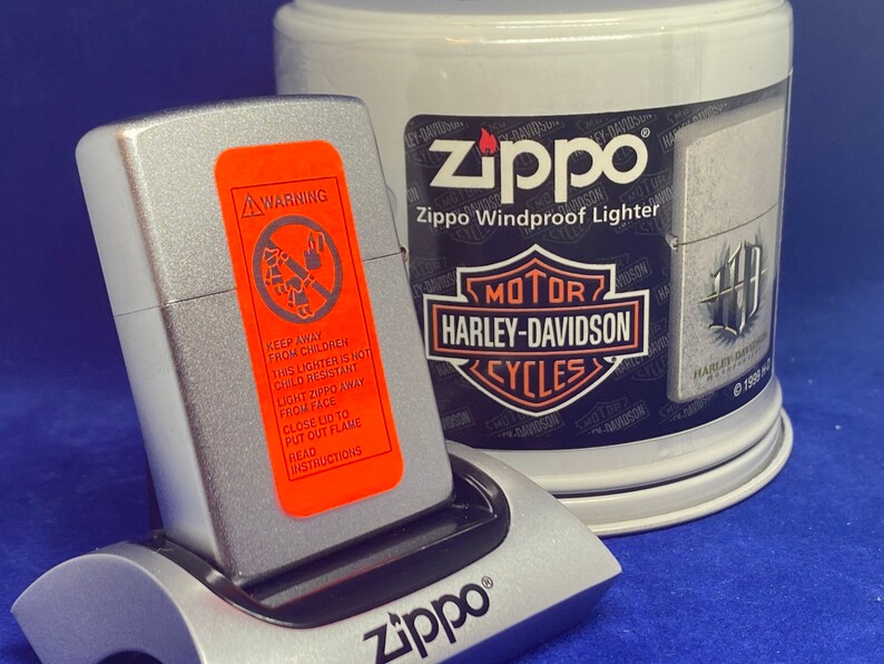 Puede incluir: Un encendedor Zippo plateado con una etiqueta de advertencia naranja y un soporte de carga plateado. La etiqueta dice "Mantener alejado de los ni&ntilde;os" y otras instrucciones de seguridad. Un recipiente blanco con la marca Harley-Davidson est&aacute; en el fondo.