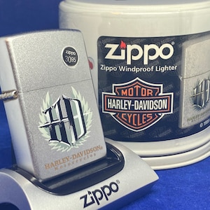Puede incluir: Un encendedor Zippo plateado con un emblema de Harley-Davidson Motorcycles, exhibido en un soporte plateado. El encendedor tiene un acabado texturizado y un logotipo en blanco y negro. Un contenedor Zippo Windproof Lighter est&aacute; en el fondo.
