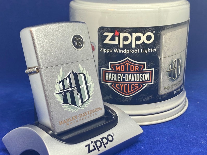 Puede incluir: Un encendedor Zippo plateado con un emblema de Harley-Davidson Motorcycles, exhibido en un soporte plateado. El encendedor tiene un acabado texturizado y un logotipo en blanco y negro. Un contenedor Zippo Windproof Lighter est&aacute; en el fondo.