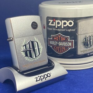 Puede incluir: Un encendedor Zippo plateado a prueba de viento con el logotipo de Harley-Davidson, colocado en un soporte plateado. El encendedor y el soporte est&aacute;n sobre una superficie azul. Una lata Zippo con gr&aacute;ficos de Harley-Davidson es visible.