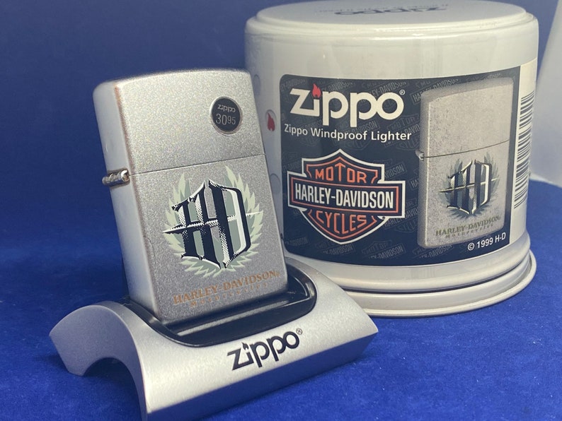 Puede incluir: Un encendedor Zippo plateado a prueba de viento con el logotipo de Harley-Davidson, colocado en un soporte plateado. El encendedor y el soporte est&aacute;n sobre una superficie azul. Una lata Zippo con gr&aacute;ficos de Harley-Davidson es visible.