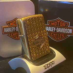 May include: A brass-toned metal lighter featuring a raised diamond plate texture and a Harley-Davidson logo. The lighter rests on a silver Zippo display stand in front of a matching silver tin and a black box, both displaying the Harley-Davidson logo.