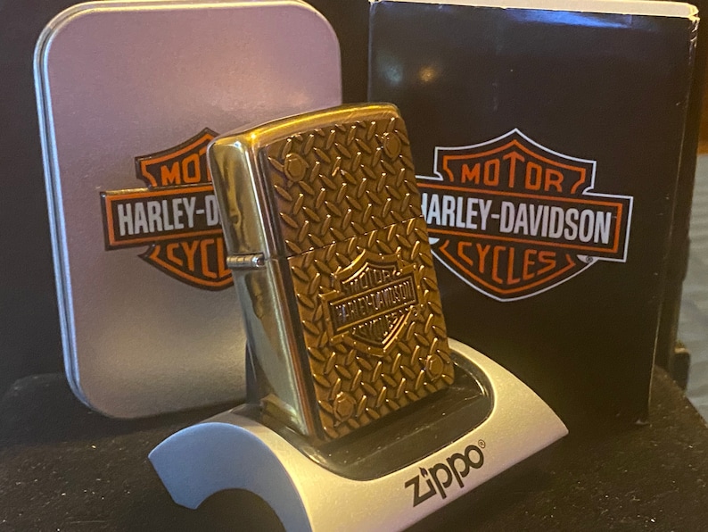May include: A brass-toned metal lighter featuring a raised diamond plate texture and a Harley-Davidson logo. The lighter rests on a silver Zippo display stand in front of a matching silver tin and a black box, both displaying the Harley-Davidson logo.