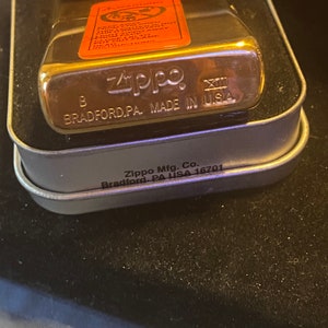 May include: A gold-tone Zippo lighter with a metallic finish, housed in a metal case. The lighter features an orange warning label on the front and the text Zippo, B, XIII, and MADE IN U.S.A. BRADFORD, PA. The case is marked Zippo Mfg. Co. Bradford, PA USA 16701.