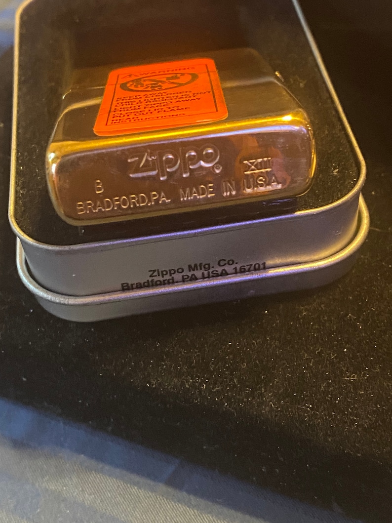 May include: A gold-tone Zippo lighter with a metallic finish, housed in a metal case. The lighter features an orange warning label on the front and the text Zippo, B, XIII, and MADE IN U.S.A. BRADFORD, PA. The case is marked Zippo Mfg. Co. Bradford, PA USA 16701.
