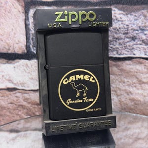 May include: A black Zippo lighter with a Camel logo, featuring a camel silhouette and the text "Genuine Taste." The lighter is in its original display case, which reads "Lifetime Guarantee."