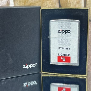 Könnte beinhalten: Ein silberner Zippo-Feuerzeug in einer schwarzen Box. Das Feuerzeug hat ein weißes Design mit dem Wort "Zippo", das mehrmals wiederholt wird, und dem Text "1977-1983 Feuerzeug".