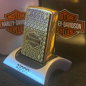 May include: A brass-toned Zippo lighter featuring a raised diamond plate texture and a Harley-Davidson logo emblem. The lighter is displayed in a silver-colored plastic stand, positioned in front of a black and orange Harley-Davidson branded box.