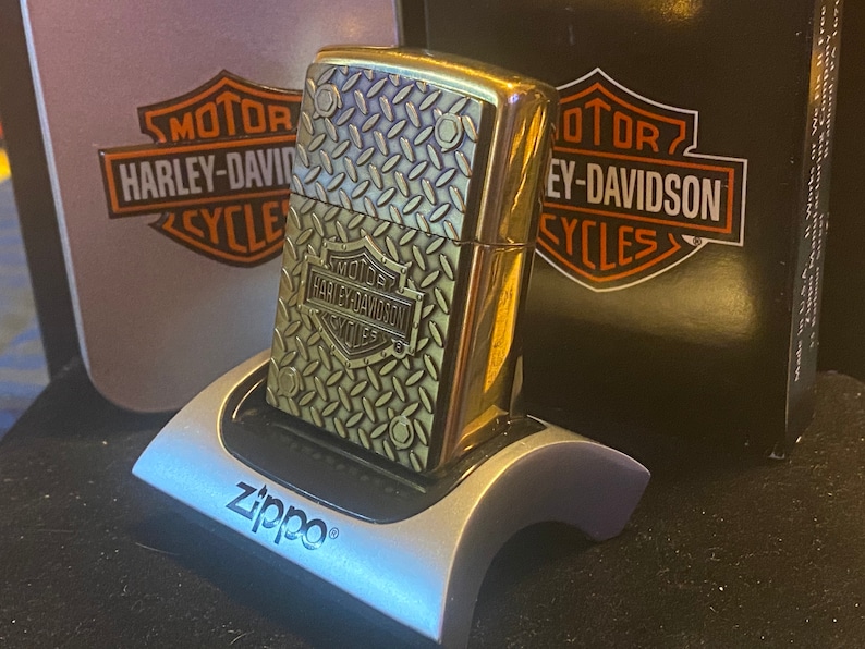 May include: A brass-toned Zippo lighter featuring a raised diamond plate texture and a Harley-Davidson logo emblem. The lighter is displayed in a silver-colored plastic stand, positioned in front of a black and orange Harley-Davidson branded box.