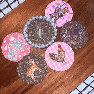 May include: A collection of decorative coasters with various designs. One coaster features a fish with a pearl border. Other coasters have a rodeo rider, a deer, a rooster, bows, and the word "MAMA". The coasters are round and placed on a wooden surface.