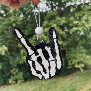 May include: Black and white air freshener in the shape of a hand making the rock and roll gesture. The hand is a skeleton design with white bones and a black glitter background. It hangs from a white string with a white bead.