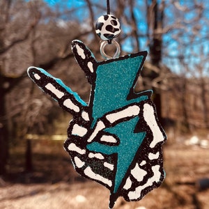 May include: A blue and white peace sign with a lightning bolt inside. The peace sign is made of glitter and has a black outline. The peace sign is hanging from a black string with a black and white bead.