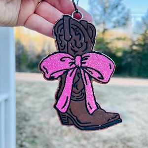 May include: A brown cowboy boot Christmas ornament with a pink glitter bow. The boot is decorated with black outlines and a glitter finish.
