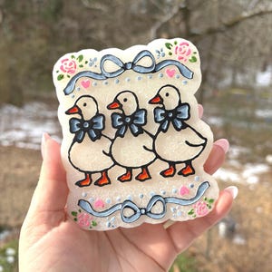 May include: A decorative, off-white candle featuring three cartoon ducks with blue bow ties and orange beaks and feet. The candle is decorated with pink roses, blue ribbons, and small pink hearts.
