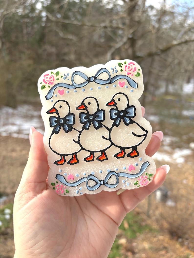 May include: A decorative, off-white candle featuring three cartoon ducks with blue bow ties and orange beaks and feet. The candle is decorated with pink roses, blue ribbons, and small pink hearts.