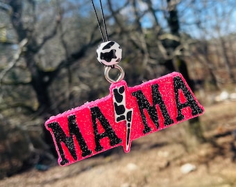 May include: A pink glittery "MAMA" car air freshener with black lettering and a lightning bolt design. It has a black string and a cow print bead. The background is blurred with trees.