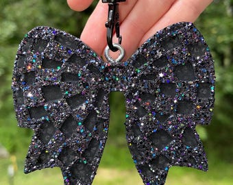 May include: A black glitter bow-shaped keychain with a black clasp and a cow-print bead. The bow has a textured pattern and is covered in iridescent glitter. The keychain is held against a blurred green background.