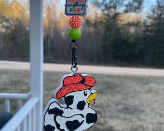 May include: A black and white cow-print duck with a red hat and a black lanyard with green and white beads. The lanyard has a charm with the text "It's fine I'm fine everything's fine".