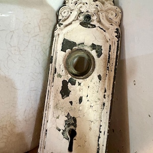 May include: A rectangular metal door plate featuring an ornate, scrolled top and a distressed white paint finish that reveals a dark base layer. The plate includes a central circular opening for a knob and a lower keyhole opening for a lock mechanism.