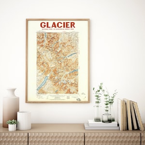 May include: A framed vintage map of Glacier National Park, US Geological Survey 1936. The map features a detailed topographical view in shades of brown, tan, and blue, with the word "GLACIER" in red at the top. The map is displayed on a light-colored wall.