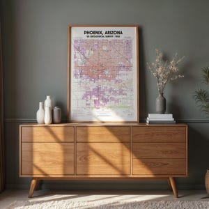May include: A framed map of Phoenix, Arizona, from the US Geological Survey, 1952, hangs above a wooden cabinet. The cabinet has six drawers and is made of light-colored wood. Decorative vases and a small plant are on the cabinet.