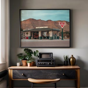 May include: A framed photograph of a vintage Conoco gas station in a desert landscape. The image is displayed above a wooden console table with a small black radio, a vintage television, plants in terracotta pots, and a gold vase.