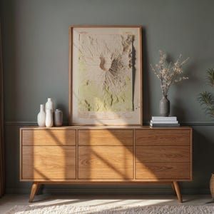 May include: A mid-century modern wooden dresser with six drawers and tapered legs. A framed topographical map hangs above the dresser. Decorative vases, books, and a potted plant complete the room's decor. The room has a neutral color palette.