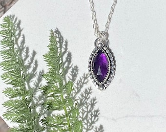 May include: A silver chain necklace with a purple amethyst gemstone pendant. The pendant is set in a silver bezel with a beaded edge.