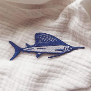 Può includere: Una toppa ricamata di un pesce marlin blu e bianco. Il marlin è rivolto a destra e ha un lungo becco appuntito.
