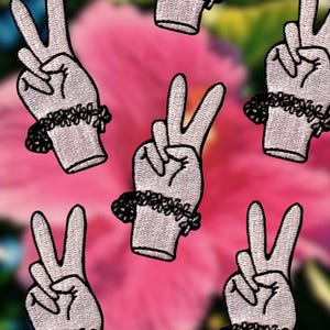 May include: Embroidered patches featuring a hand making the peace sign. The hands are white with black outlines and a black floral wristband. The background is a blurred pink and green floral pattern.