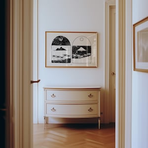 May include: A white chest of drawers with two drawers and gold accents sits in a room with white walls and a framed print of two black and white illustrations. The print depicts two islands with palm trees and a crescent moon or sun above each.
