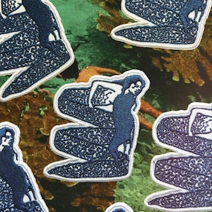 Könnte beinhalten: Gestickte Aufnäher mit einer stilisierten dunkelblauen Figur und weißem Rand. Die Figur sitzt auf einer strukturierten, wellenartigen Form. Die Aufnäher sind vor einem grün-braunen Hintergrund platziert.