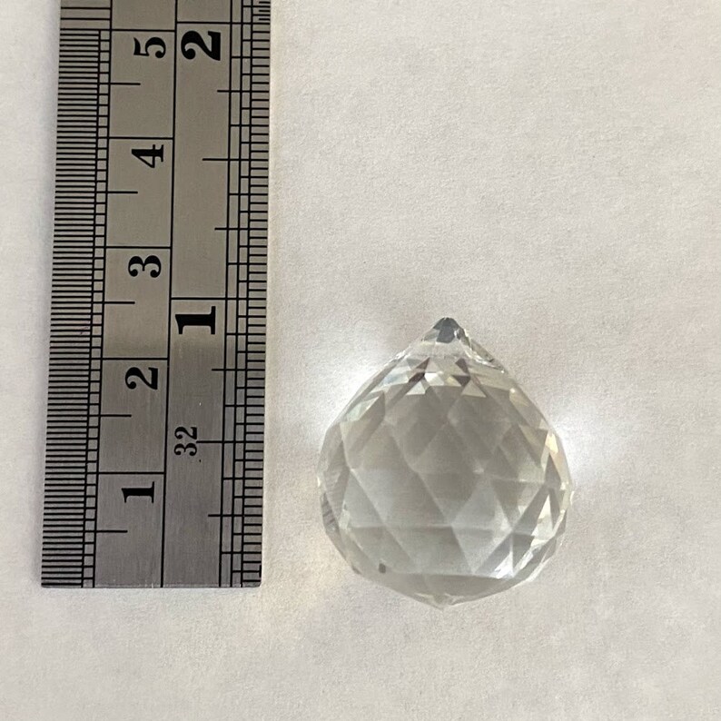 May include: A clear glass crystal prism pendant, approximately 1 inch in diameter, with a faceted design. The pendant is shown next to a ruler for size reference.