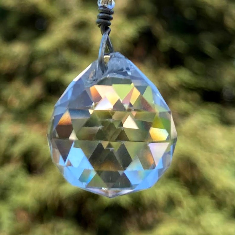 May include: A clear crystal prism with a faceted design hangs from a silver wire. The prism reflects light, creating a rainbow effect.