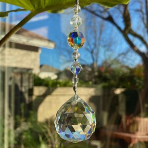 May include: A clear crystal suncatcher with a faceted, spherical bottom and a rainbow-colored, multi-faceted center bead. The suncatcher is suspended by a thin, silver-colored wire and clear beads. The background shows a blurred outdoor scene with a house and trees.
