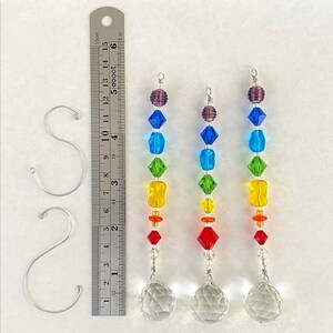 May include: Three rainbow crystal suncatchers with clear crystal balls at the bottom. Each suncatcher has a purple, blue, green, yellow, orange, and red crystal bead. The suncatchers are hanging from silver hooks. A ruler is in the background for size reference.