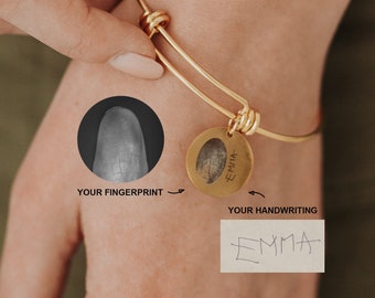 Puede incluir: Un brazalete de oro con un dije que presenta una huella digital y el nombre escrito a mano "EMMA".