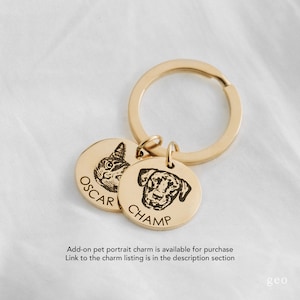 May include: Gold-toned keyring with two round charms. One charm features a black and white engraved image of a cat with the name "OSCAR" engraved below. The other charm features a black and white engraved image of a dog with the name "CHAMP" engraved below.