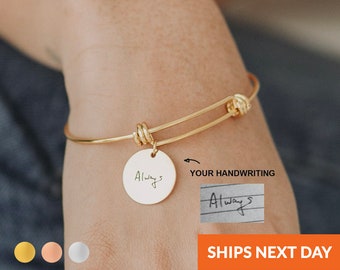 Könnte beinhalten: Goldenes Armreif-Armband mit einem runden Anhänger, auf dem "Always" in kursivschrift steht. Der Anhänger ist mit einem Knoten am Armband befestigt. Das Armband wird am Handgelenk einer Person gezeigt.