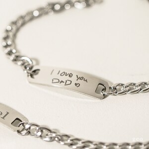 Puede incluir: Dos pulseras de cadena plateadas con una etiqueta rectangular. La etiqueta de una pulsera tiene el mensaje escrito a mano "Te quiero pap&aacute;" con un coraz&oacute;n dibujado al lado.