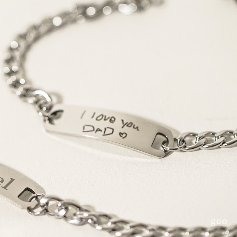 Puede incluir: Dos pulseras de cadena plateadas con una etiqueta rectangular. La etiqueta de una pulsera tiene el mensaje escrito a mano "Te quiero pap&aacute;" con un coraz&oacute;n dibujado al lado.