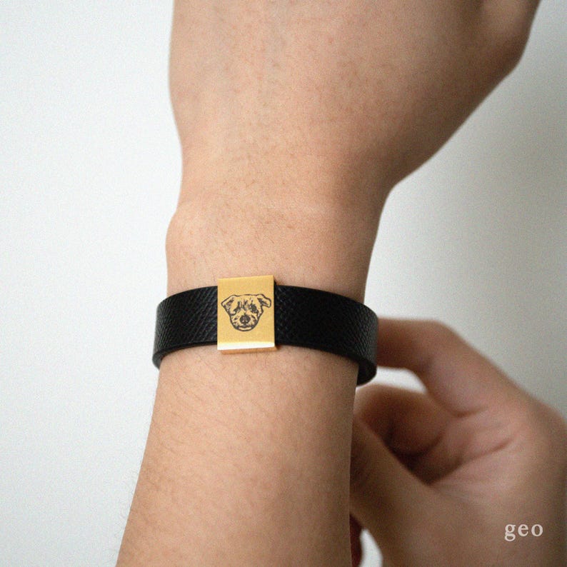 Puede incluir: Una pulsera de cuero negro con un dije cuadrado dorado con la cara de un perro.