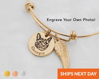 Könnte beinhalten: Goldenes Armreif-Armband mit einem runden Anhänger, der mit einem Katzenfoto und dem Namen "Georgie" graviert ist. Der Anhänger ist an einem kleinen goldenen Flügel-Anhänger befestigt.  Gravieren Sie Ihr eigenes Foto! Versand am nächsten Tag.