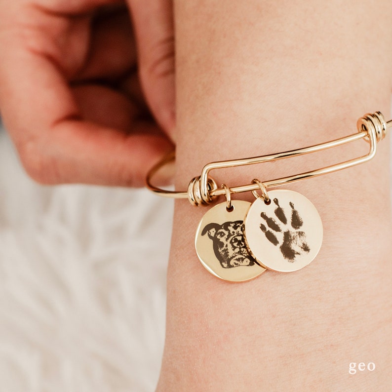 May include: A gold expandable bracelet with two charms. One charm features a black and white photo of a dog's face. The other charm features a black paw print.