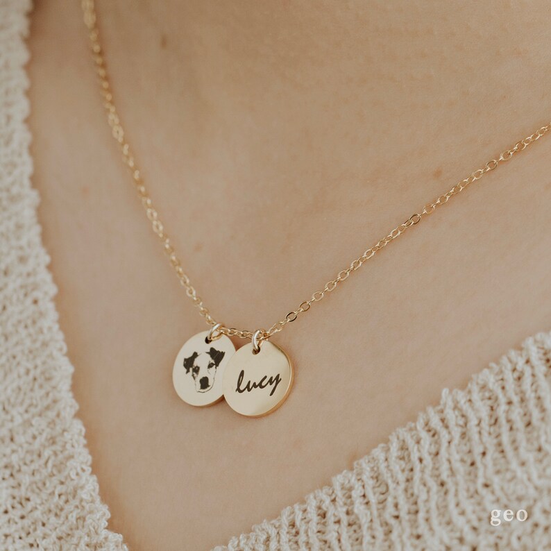 Op de afbeelding: Gouden ketting met twee ronde hangers. Een hanger heeft een zwart-wit foto van een hond, de andere hanger heeft de naam "Lucy" gegraveerd.