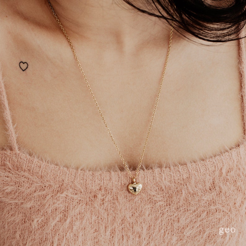 May include: A gold chain necklace with a small, heart-shaped pendant. The pendant has a textured surface.
