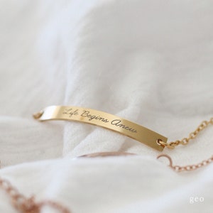 May include: Gold bar necklace with a delicate chain. The bar has the inscription "Life Begins Anew".