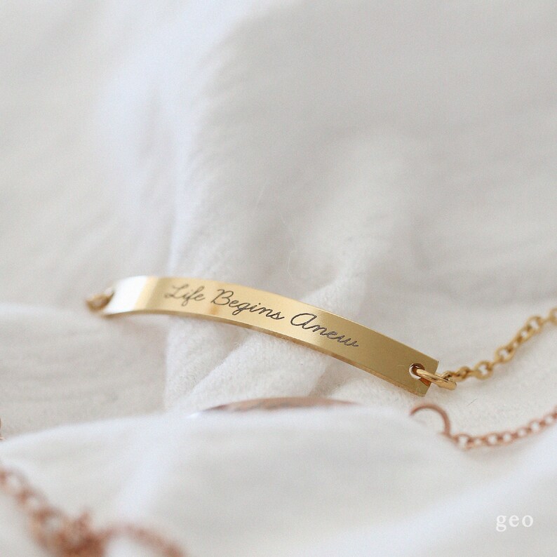 May include: Gold bar necklace with a delicate chain. The bar has the inscription "Life Begins Anew".